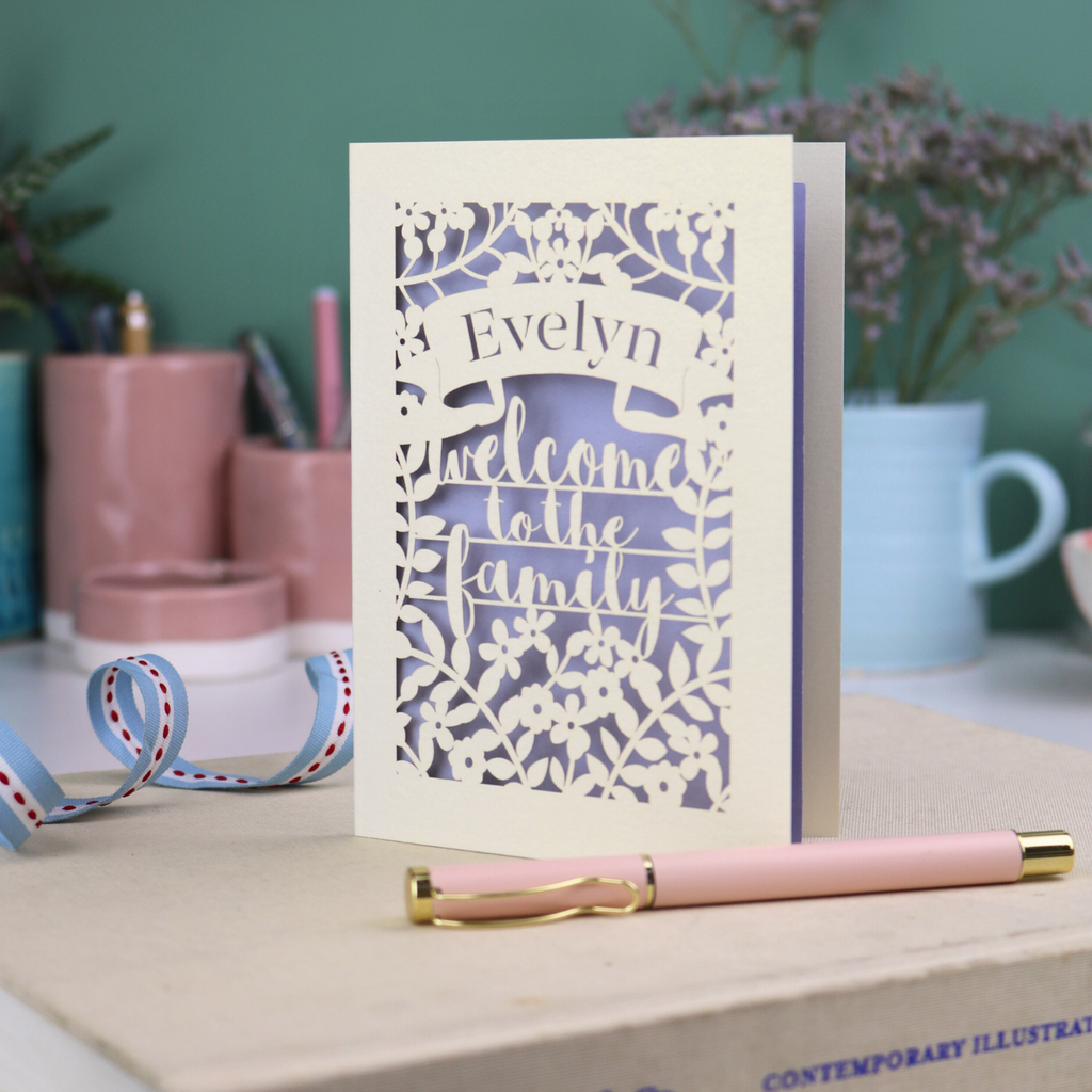 A floral laser cut wedding card personalised with a name and the text "Welcome to the Family", standing on a desk with stationery items.