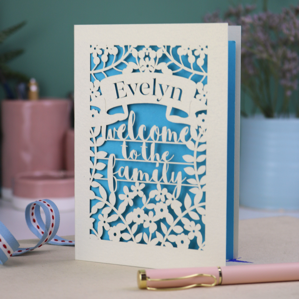 A floral laser cut wedding card personalised with a name and the text "Welcome to the Family", standing on a desk with stationery items.