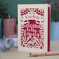A floral laser cut adoption card personalised with a name and the text "Welcome to the Family", standing on a desk with stationery items.