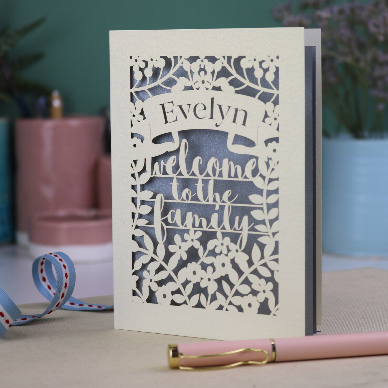 A floral laser cut adoption card personalised with a name and the text "Welcome to the Family", standing on a desk with stationery items.