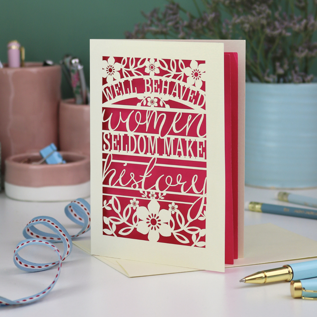 Decorative International Women's Day card with text 'Well Behaved Women Seldom Make History' and floral design on a desk with stationery items.