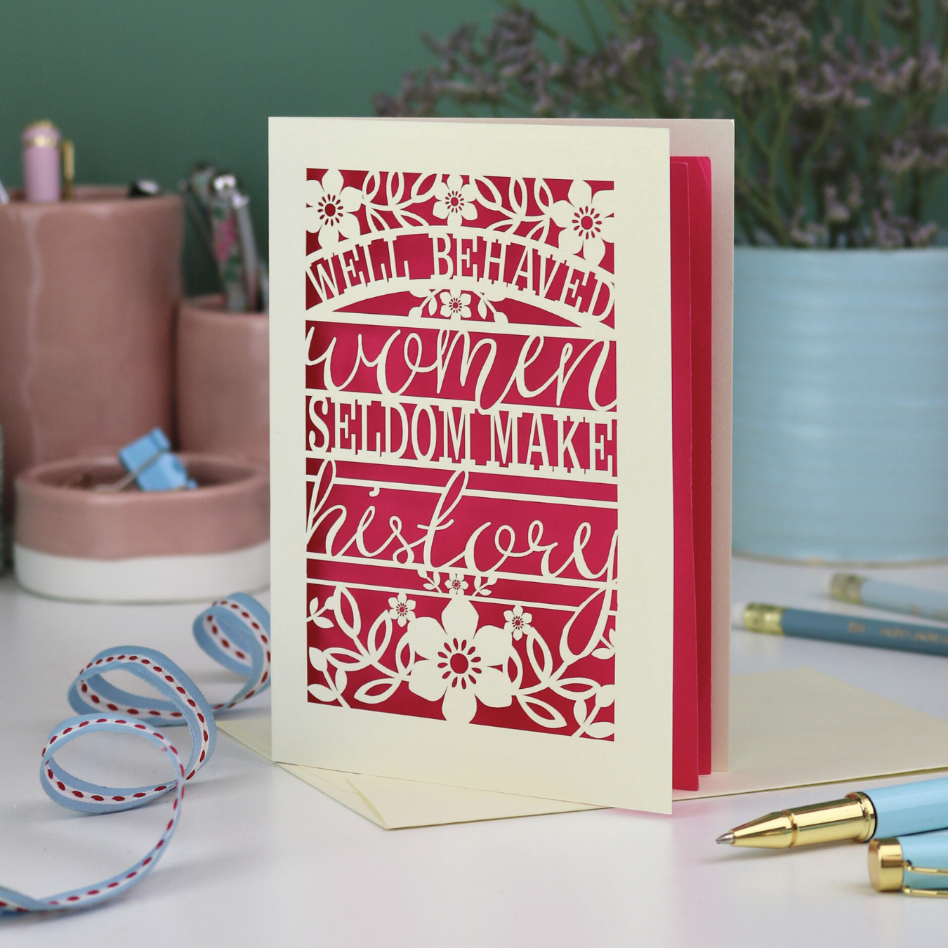 Decorative International Women's Day card with text 'Well Behaved Women Seldom Make History' and floral design on a desk with stationery items.
