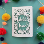 A personalised laser cut achievement card that says "Well Done" and has the recipient's name in a banner