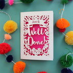 A personalised cut out celebration card that says "Well Done" and has the recipient's name in a banner
