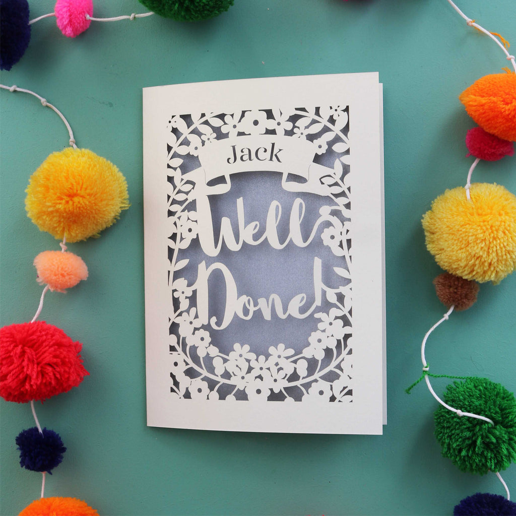 A personalised laser cut celebration card that says "Well Done" and has the recipient's name in a banner
