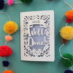 A personalised laser cut celebration card that says "Well Done" and has the recipient's name in a banner
