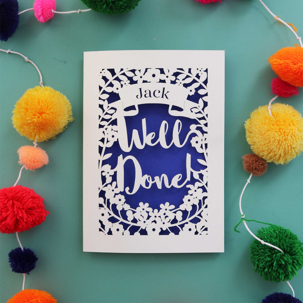 A personalised congrats laser cut card that says "Well Done" and has the recipient's name in a banner