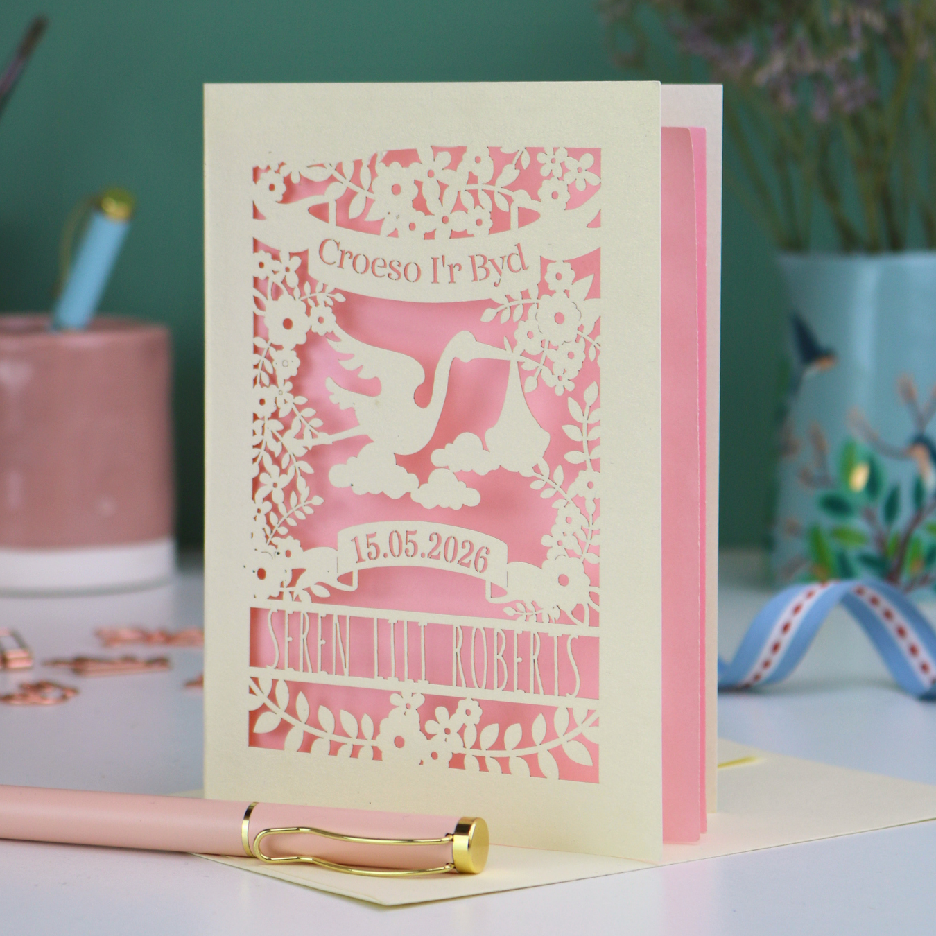 Personalised laser-cut Welsh Baby card with stork design and text: "Croeso I'r Byd", personalised with the date of birth and baby's name, on a blurred background