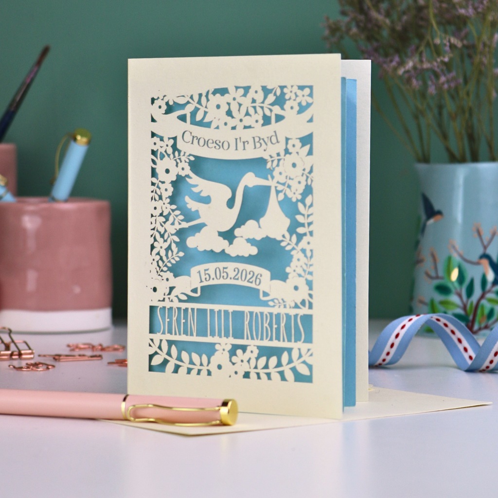 Personalised cut out Welsh New Baby card with stork design and text: "Croeso I'r Byd", personalised with the date of birth and baby's name, on a blurred background