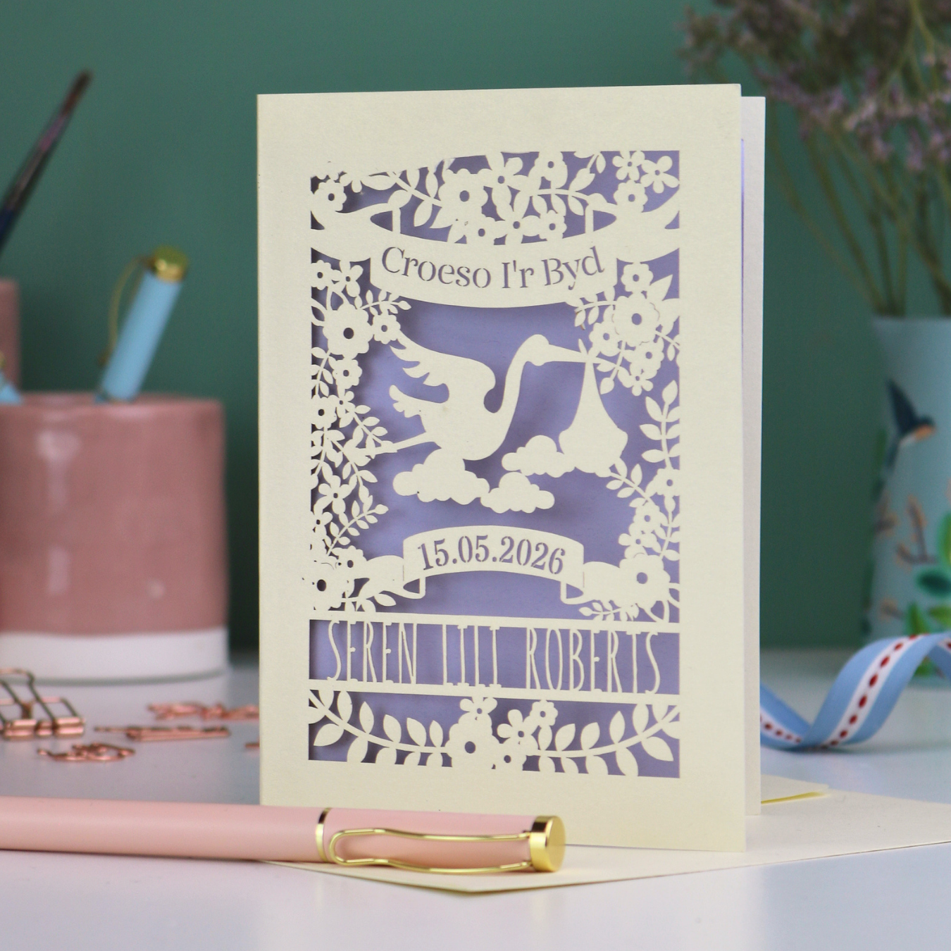 Personalised paper-cut Welsh New Baby card with stork design and text: "Croeso I'r Byd", personalised with the date of birth and baby's name, on a blurred background