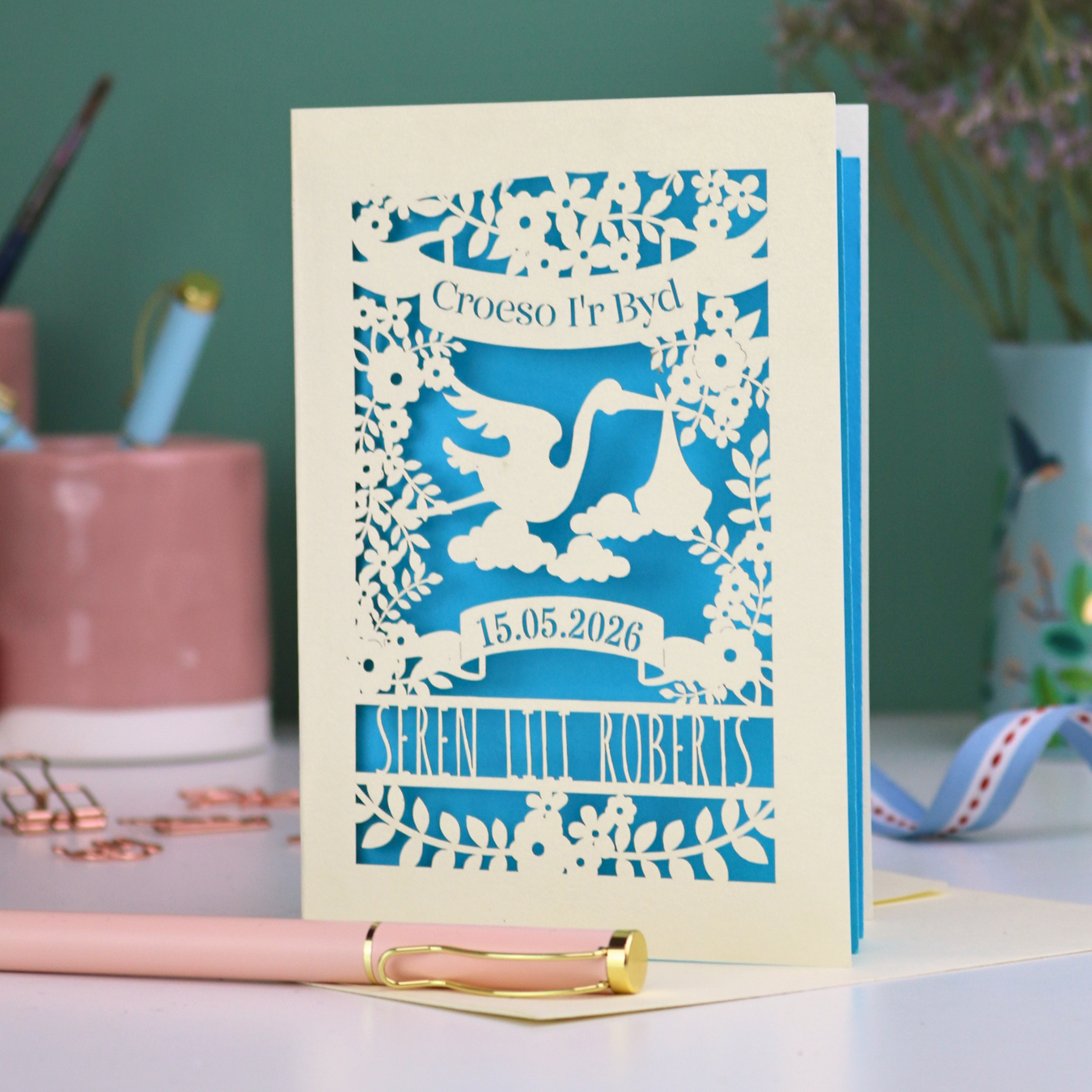 Personalised Welsh New Baby card with stork design and text: "Croeso I'r Byd", personalised with the date of birth and baby's name, on a blurred background