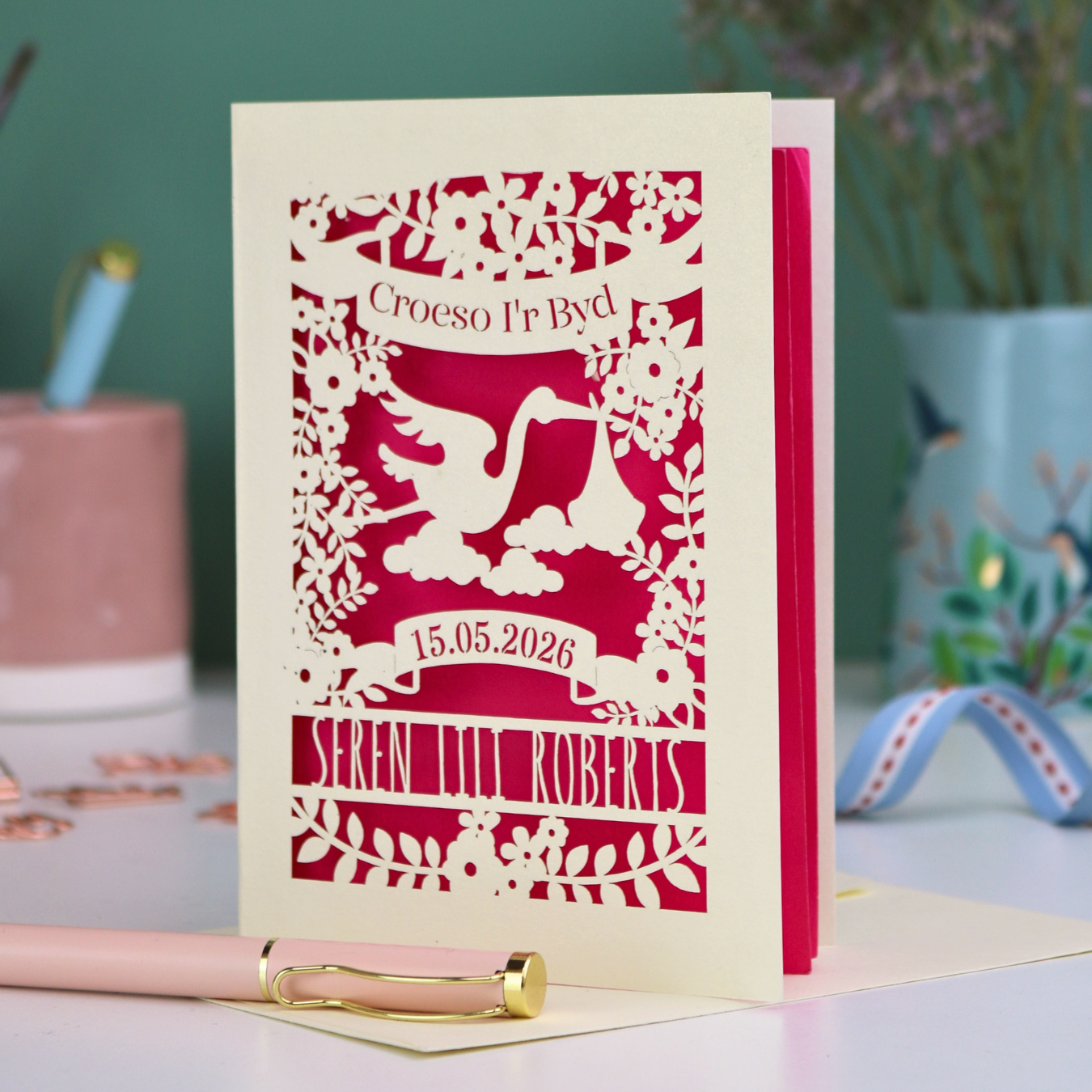 Personalized laser-cut Welsh New Baby card with stork design and text: "Croeso I'r Byd", personalised with the date of birth and baby's name, on a blurred background