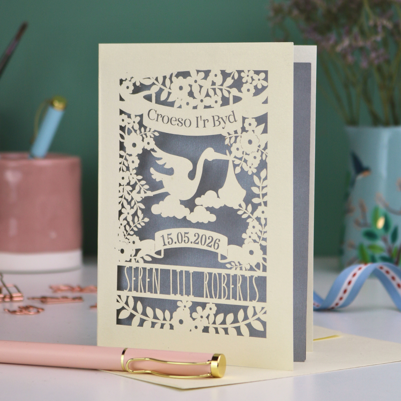 Personalized laser-cut Welsh New Baby card with stork design and text: "Croeso I'r Byd", personalised with the date of birth and baby's name, on a blurred background
