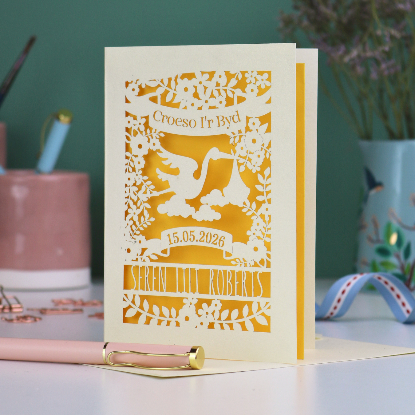 Personalized laser-cut Welsh New Baby card with stork design and text: "Croeso I'r Byd", personalised with the date of birth and baby's name, on a blurred background