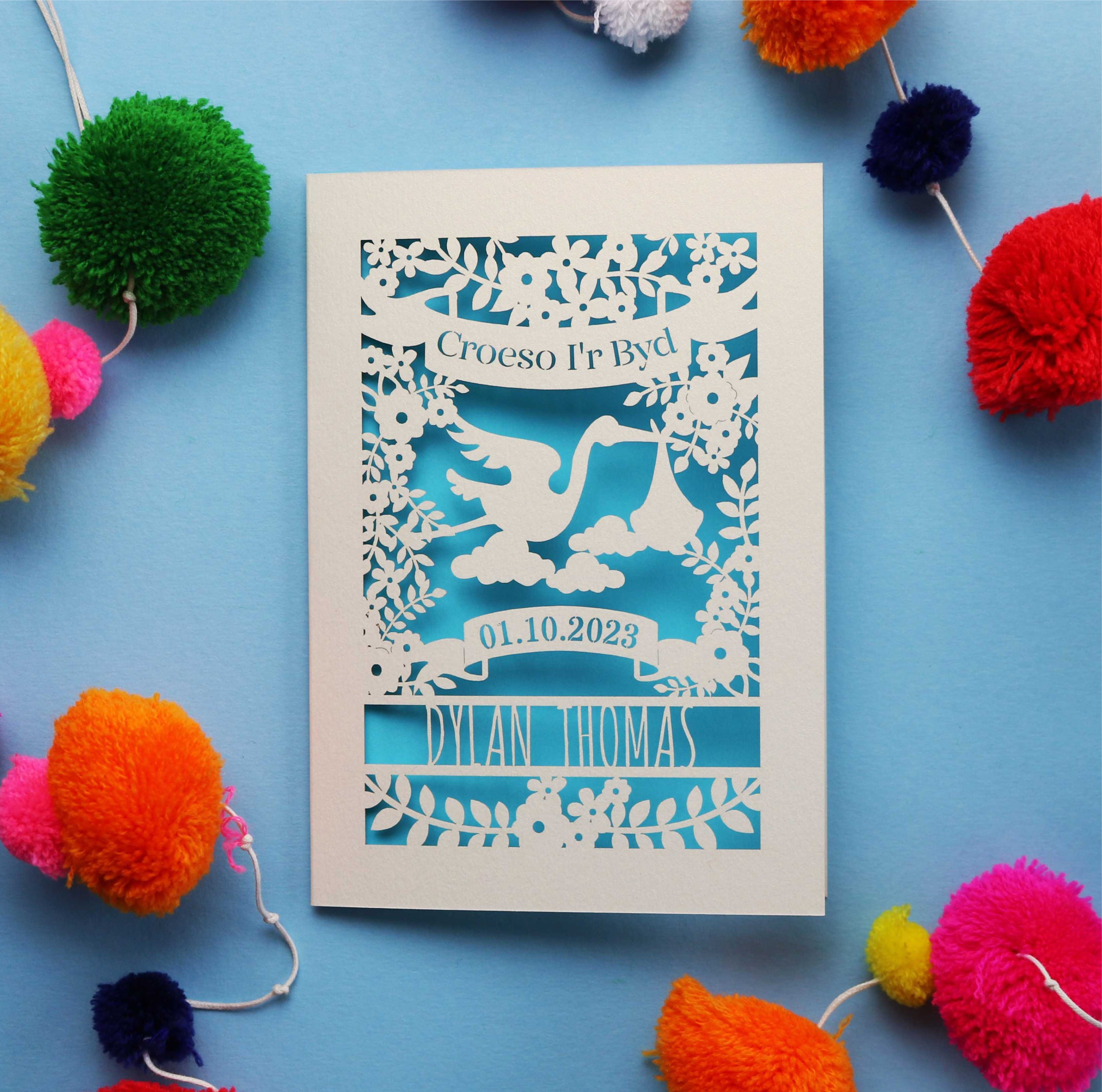 Welcome to the world in Welsh, personalised new baby card.  Showing a lovely stork carrying a baby, surrounded with flowers and leaves.  Top banner states "Croeso i'r Byd.  Personalise with baby's name and date of birth. Shown  lasercut from cream card and finished with a peacock blue insert paper.