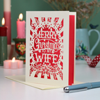 A laser-cut Christmas greeting card with the text 'MERRY Christmas TO MY WIFE' stands open on a cream envelope on a white table. The background features a softly blurred holiday scene with lit pink candles, a green wall, and a blue pen in the foreground.