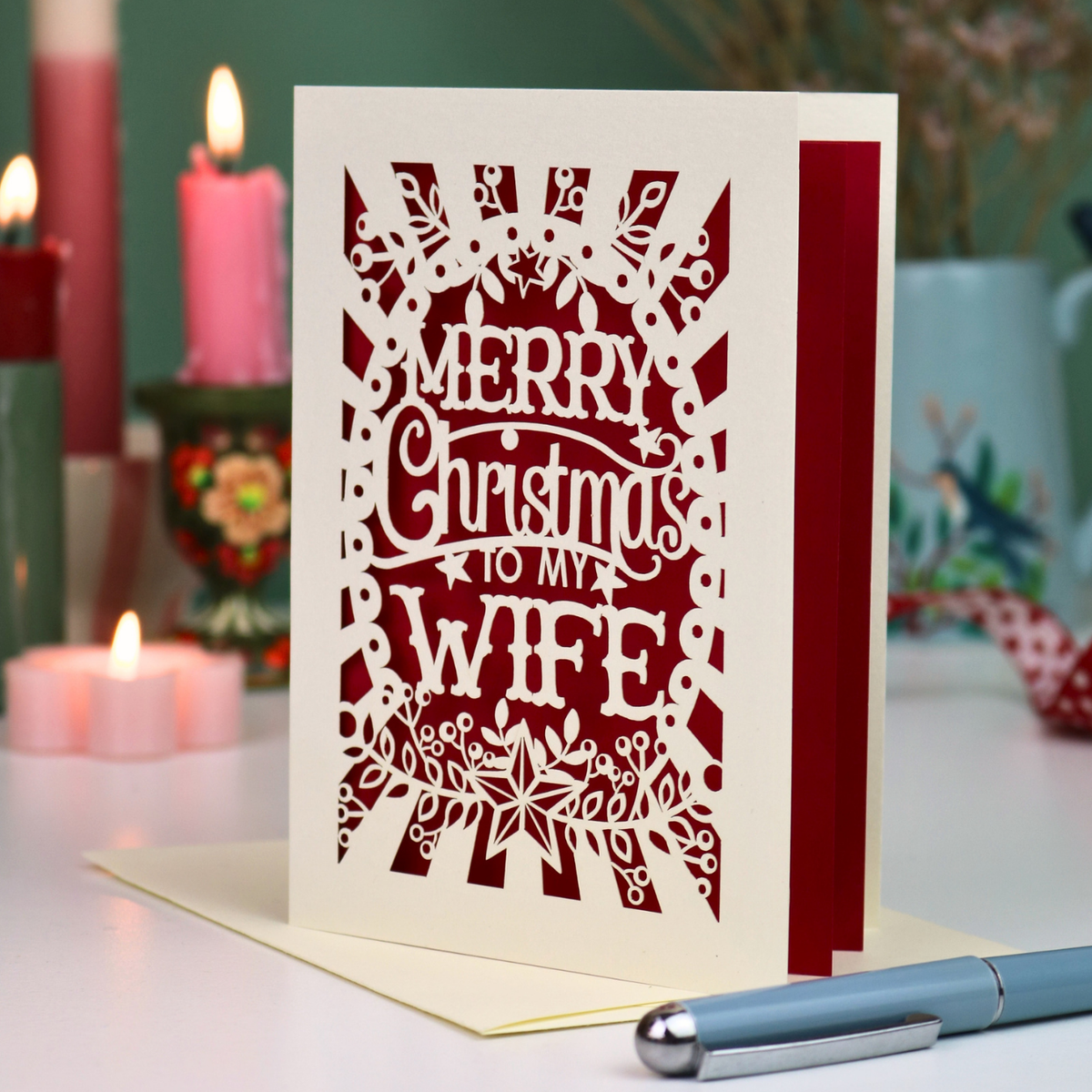 A laser-cut Christmas greeting card with the phrase 'MERRY Christmas to MY WIFE' in cut out lettering over a dark red background, standing upright on a table with candles and a pen in a softly blurred holiday setting.