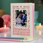 A personalized laser-cut Christmas greeting card with a photo of a couple, displayed on a table with candles and pencils in a cosy setting. Card says 'To my lovely wife Sophie at Christmas'