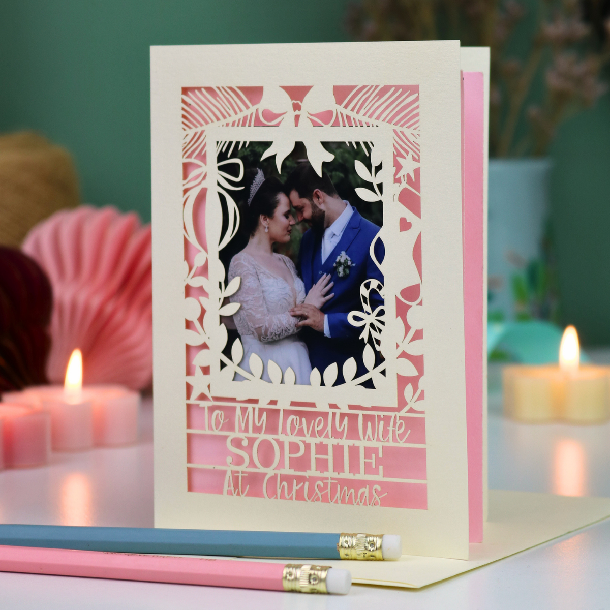 A personalized laser-cut Christmas greeting card with a photo of a couple, displayed on a table with candles and pencils in a cosy setting. Card says 'To my lovely wife Sophie at Christmas'