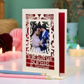 A personalized laser-cut Christmas greeting card with a photo of a couple, displayed on a table with candles and pencils in a cosy setting. Card says 'To my lovely wife Sophie at Christmas'