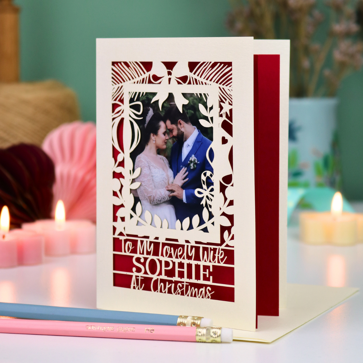 A personalized laser-cut Christmas greeting card with a photo of a couple, displayed on a table with candles and pencils in a cosy setting. Card says 'To my lovely wife Sophie at Christmas'