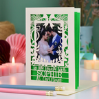 A personalised laser-cut Christmas greeting card with a photo of a couple, displayed on a table with candles and pencils in a cosy setting. Card says 'To my lovely wife Sophie at Christmas'