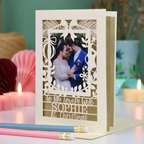 A personalized laser-cut Christmas greeting card for a wife, featuring a photo of a bride and groom and the text 'To My lovely wife SOPHIE At Christmas'. The card is displayed on a table with candles and pencils in the background.