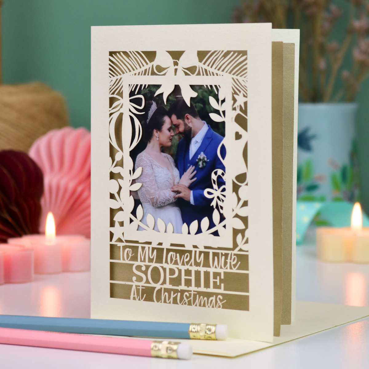 A personalized laser-cut Christmas greeting card for a wife, featuring a photo of a bride and groom and the text 'To My lovely wife SOPHIE At Christmas'. The card is displayed on a table with candles and pencils in the background.