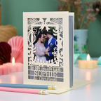 A personalized laser-cut Christmas greeting card with a photo of a couple, displayed on a table with candles and pencils in a cosy setting. Card says 'To my lovely wife Sophie at Christmas'