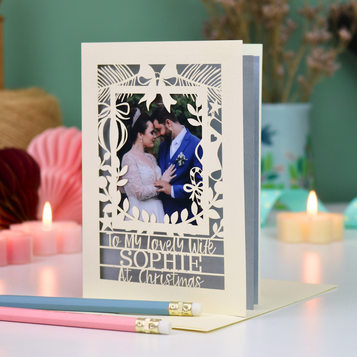 A personalized laser-cut Christmas greeting card with a photo of a couple, displayed on a table with candles and pencils in a cosy setting. Card says 'To my lovely wife Sophie at Christmas'