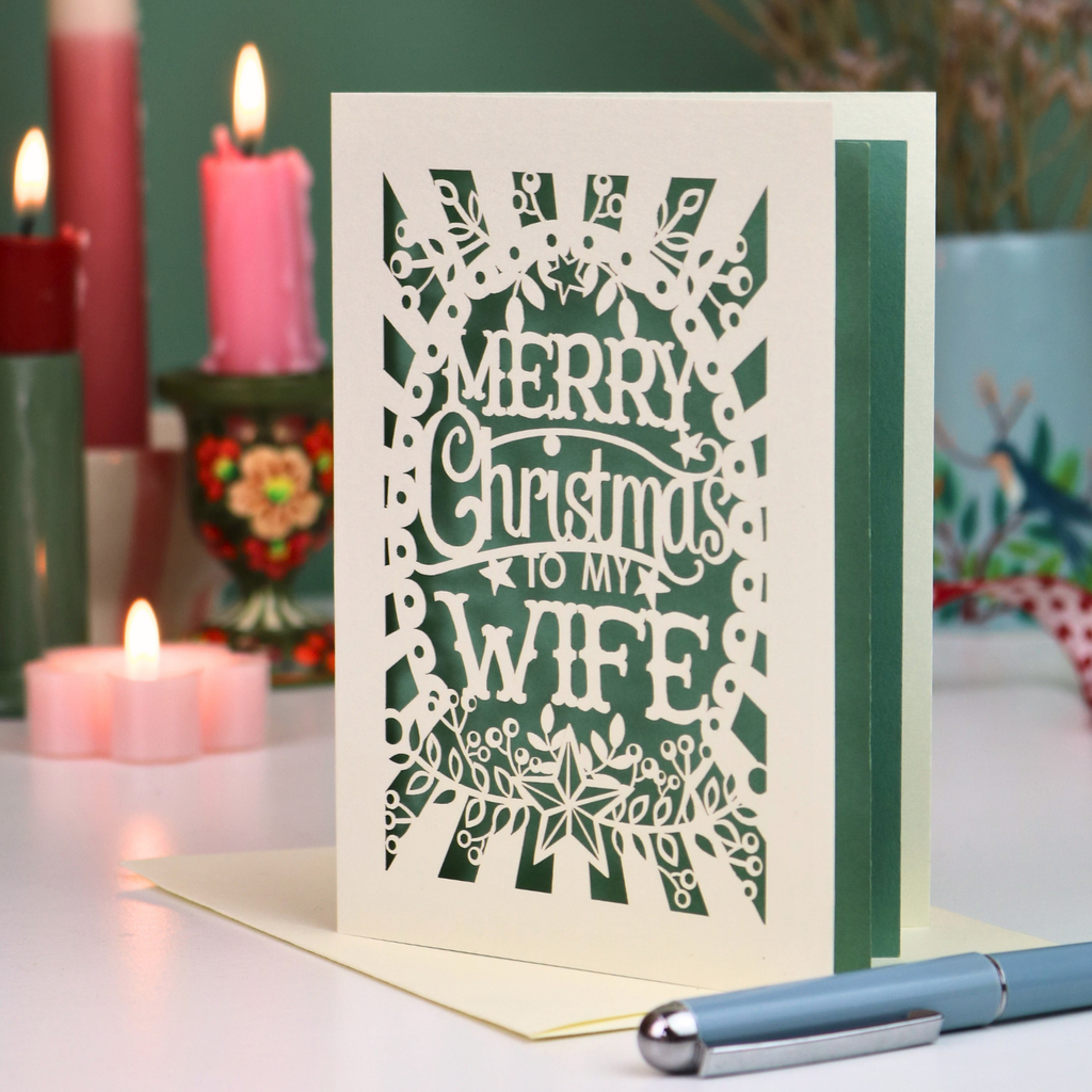 A laser-cut Christmas card with the text 'MERRY Christmas TO MY WIFE' in cut out lettering over a sage green background, displayed on a table with candles and a pen in a festive setting.