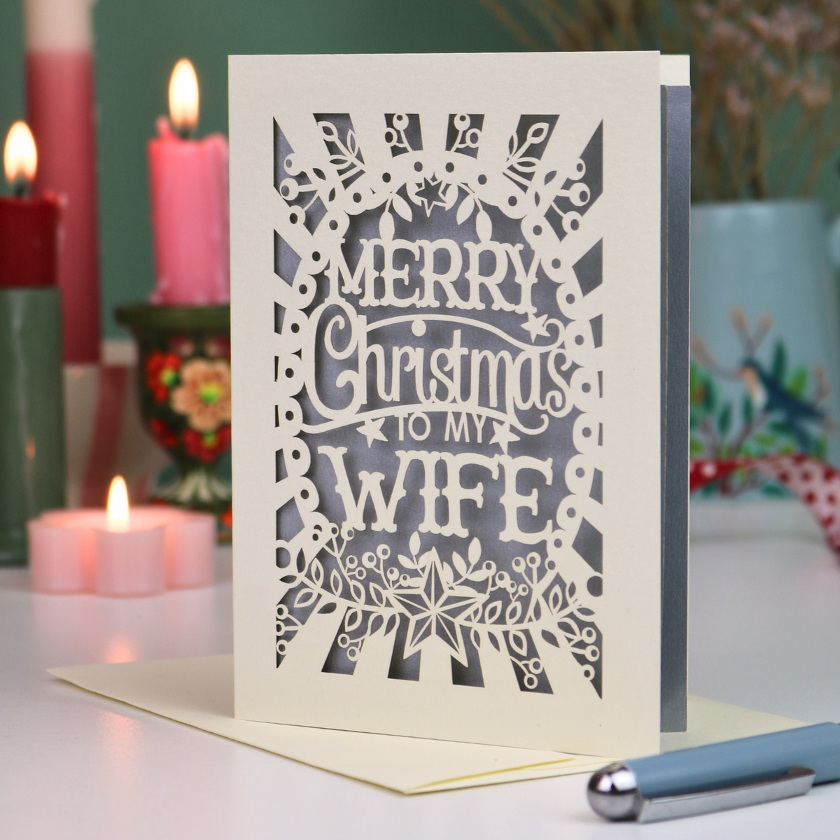 A laser-cut Christmas greeting card with the text 'MERRY Christmas TO MY WIFE' in an ornate floral border, standing upright on a table with candles and a pen blurred in the background.