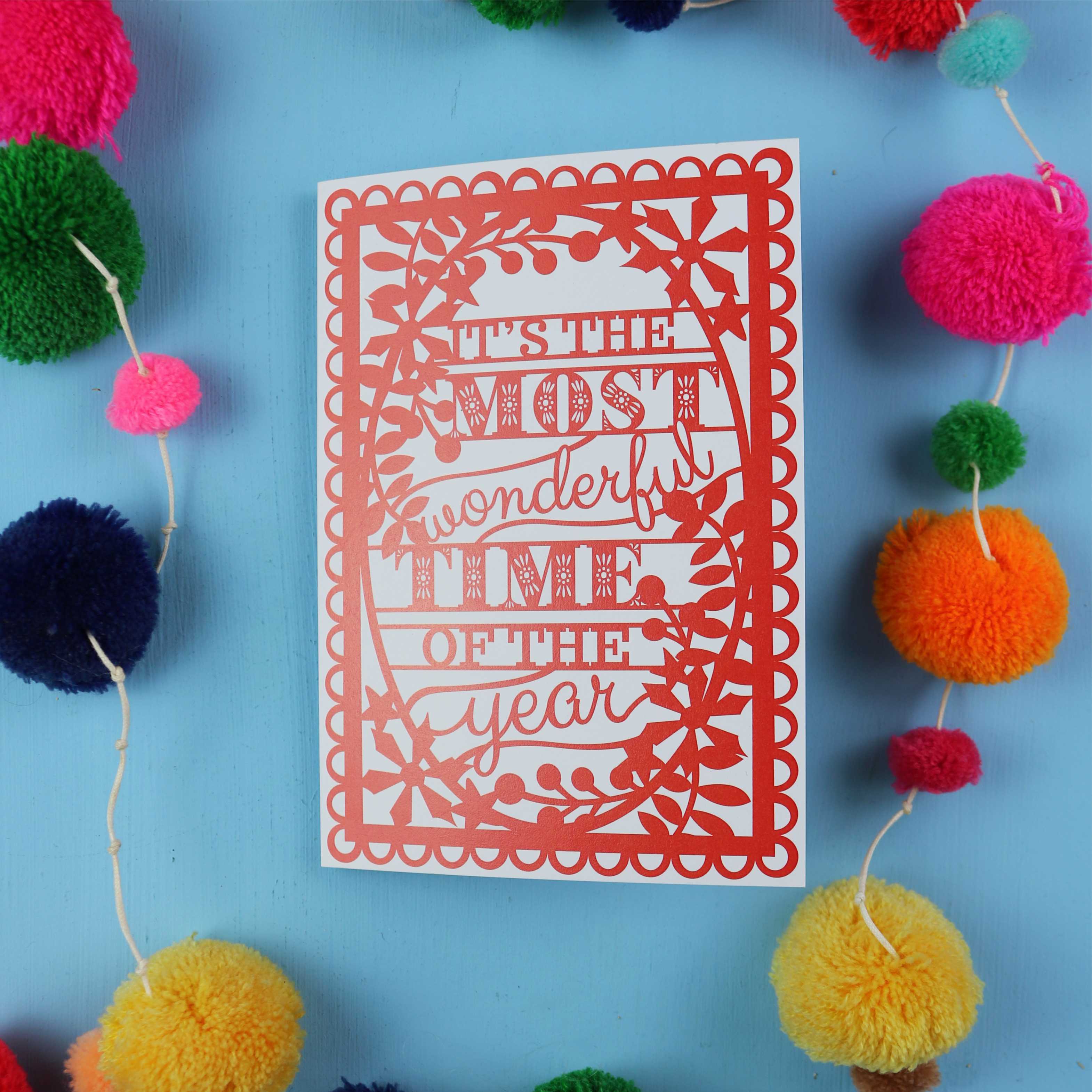 Decorative card with 'It's the most wonderful time of the year' text on a blue background with colourful pom-poms.