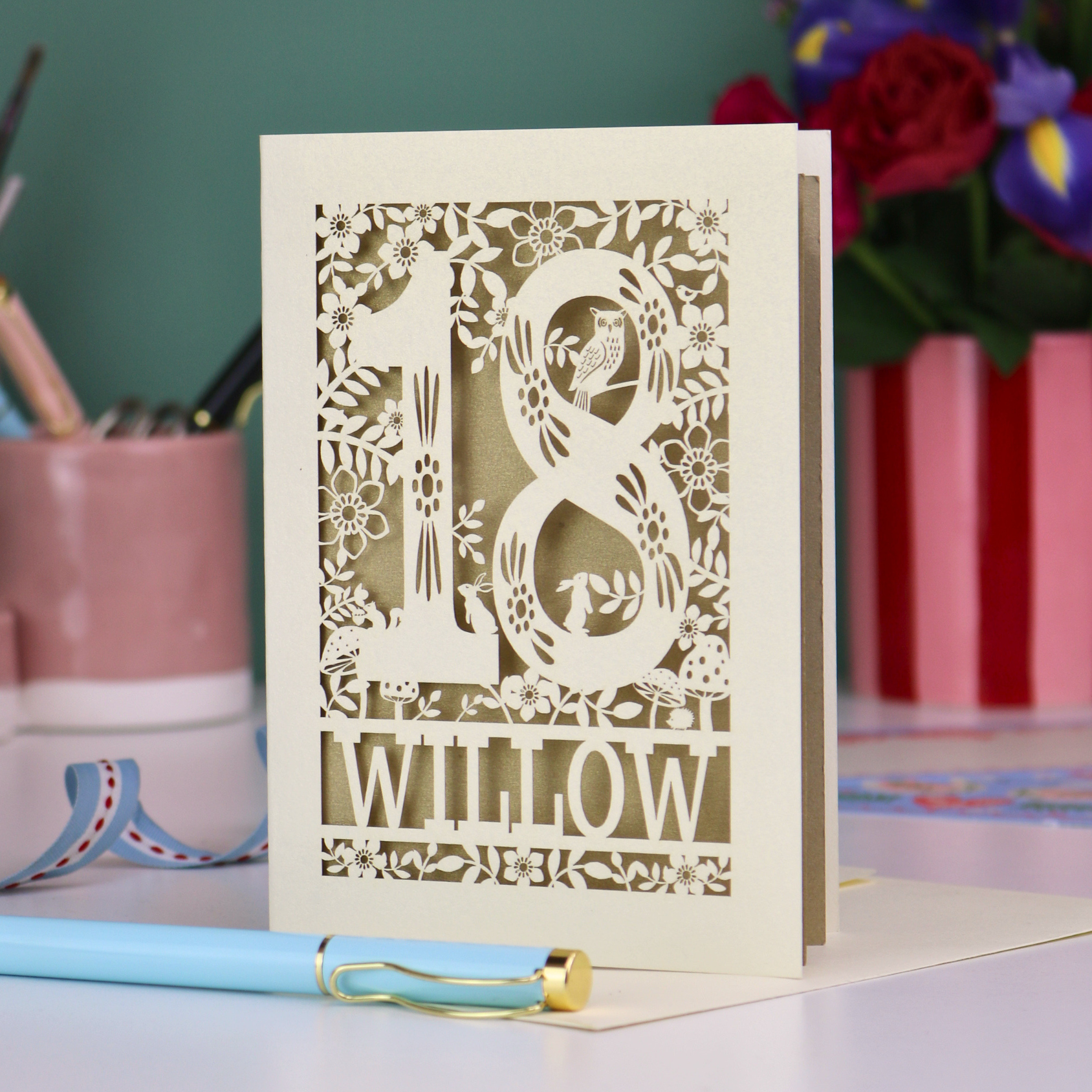Personalised laser cut 18th birthday card with '18' and personalised with the name 'Willow', featuring floral designs and woodland creatures. Card stands on a desk with stationery items.