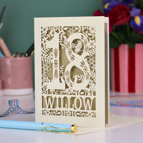 Personalised laser cut 18th birthday card with '18' and personalised with the name 'Willow', featuring floral designs and woodland creatures. Card stands on a desk with stationery items.