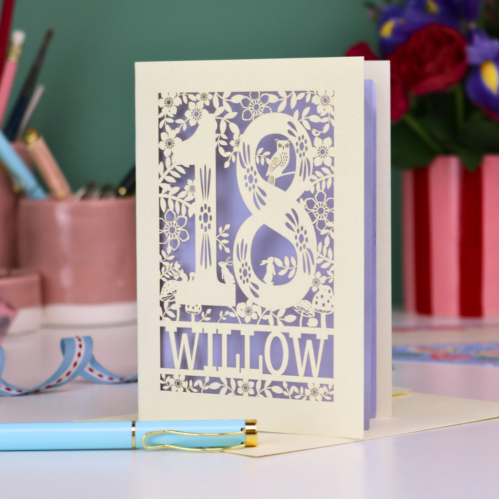 Decorative keepsake 18th birthday card with '18' and personalised with the name 'Willow', featuring floral designs and woodland creatures. Card stands on a desk with stationery items.