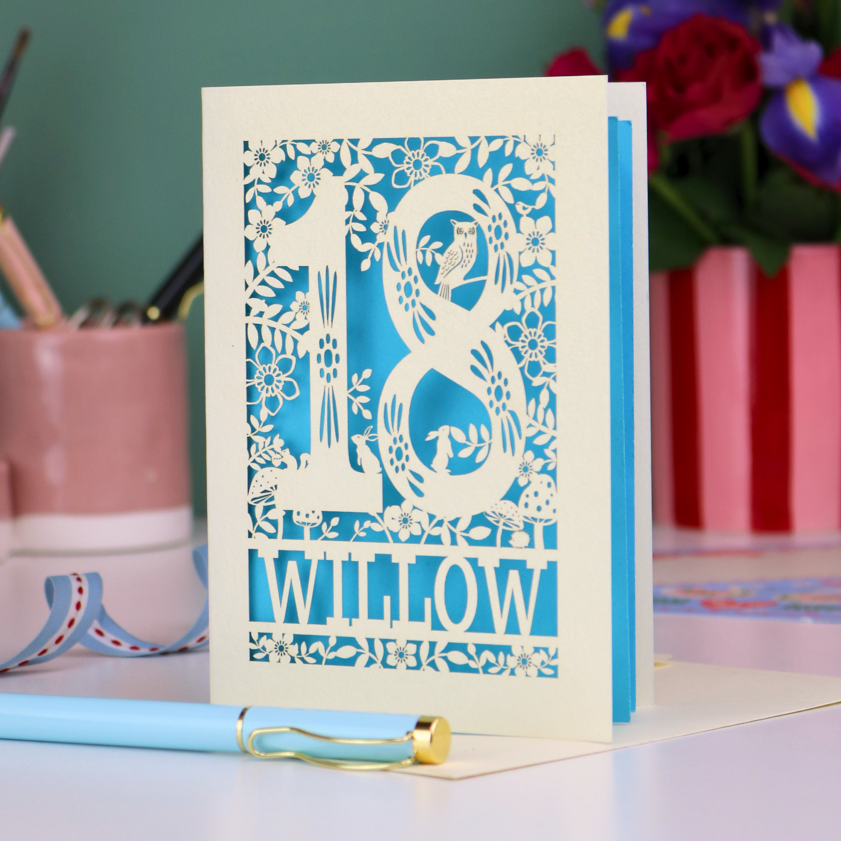 Decorative laser cut  special 18th birthday card with '18' and personalised with the name 'Willow', featuring floral designs and woodland creatures. Card stands on a desk with stationery items.