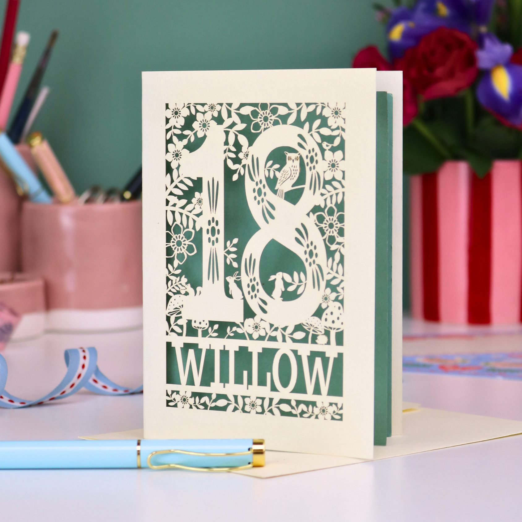 Decorative laser cut 18th birthday card with '18' and personalised with the name 'Willow', featuring floral designs and woodland creatures. Card stands on a desk with stationery items.