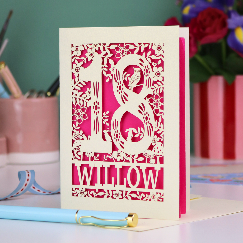 Decorative laser cut 18th birthday card with '18' and personalised with the name 'Willow', featuring floral designs and woodland creatures. Card stands on a desk with stationery items.