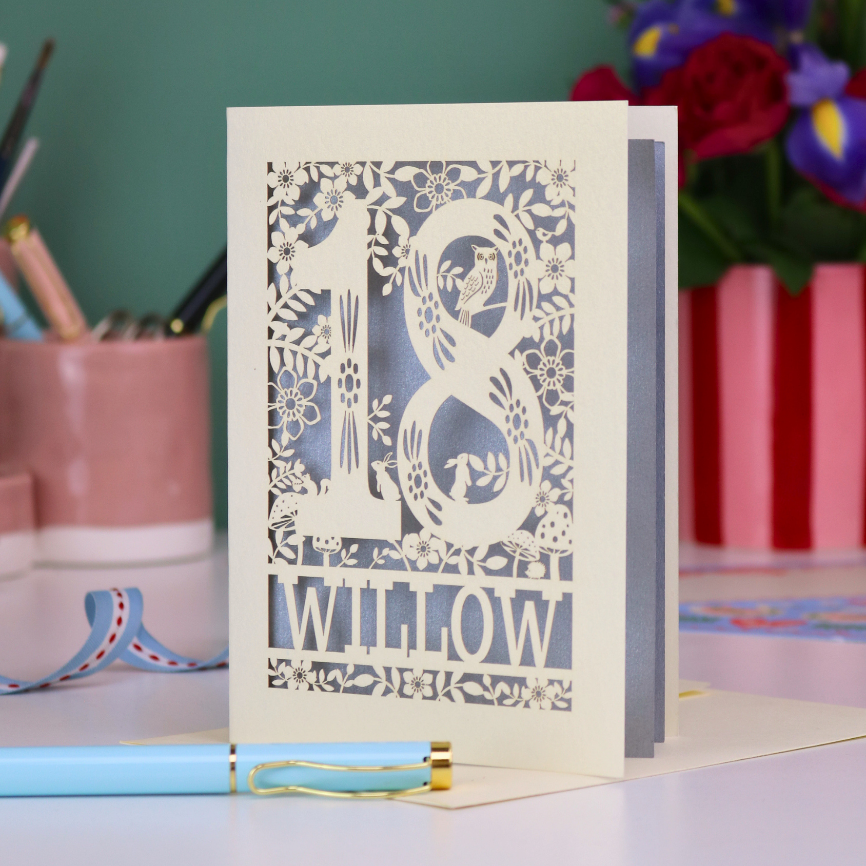 Decorative laser cut 18th birthday card with '18' and personalised with the name 'Willow', featuring floral designs and woodland creatures. Card stands on a desk with stationery items.