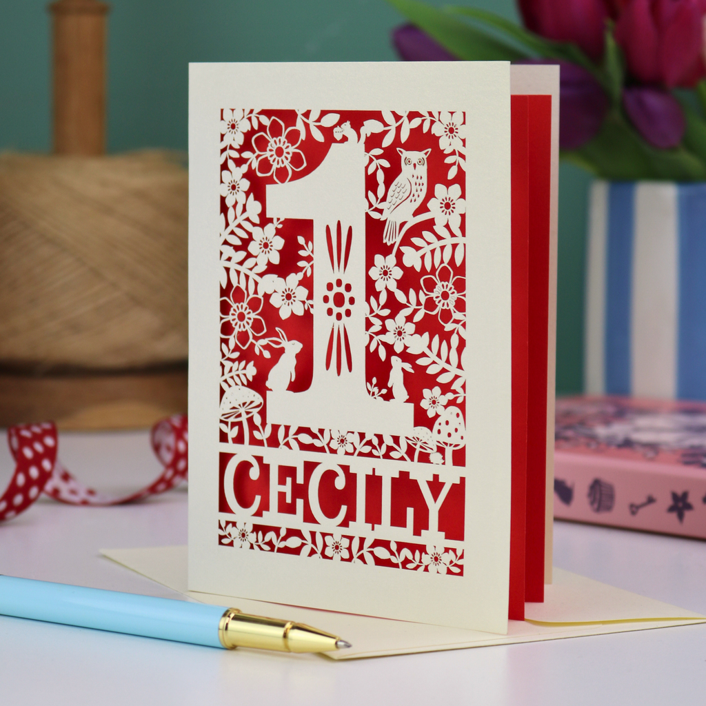 A laser cut 1st birthday card with decorative 1 and woodland design including owl, bunnies, squirrel and hedgehog, personalised with a name.