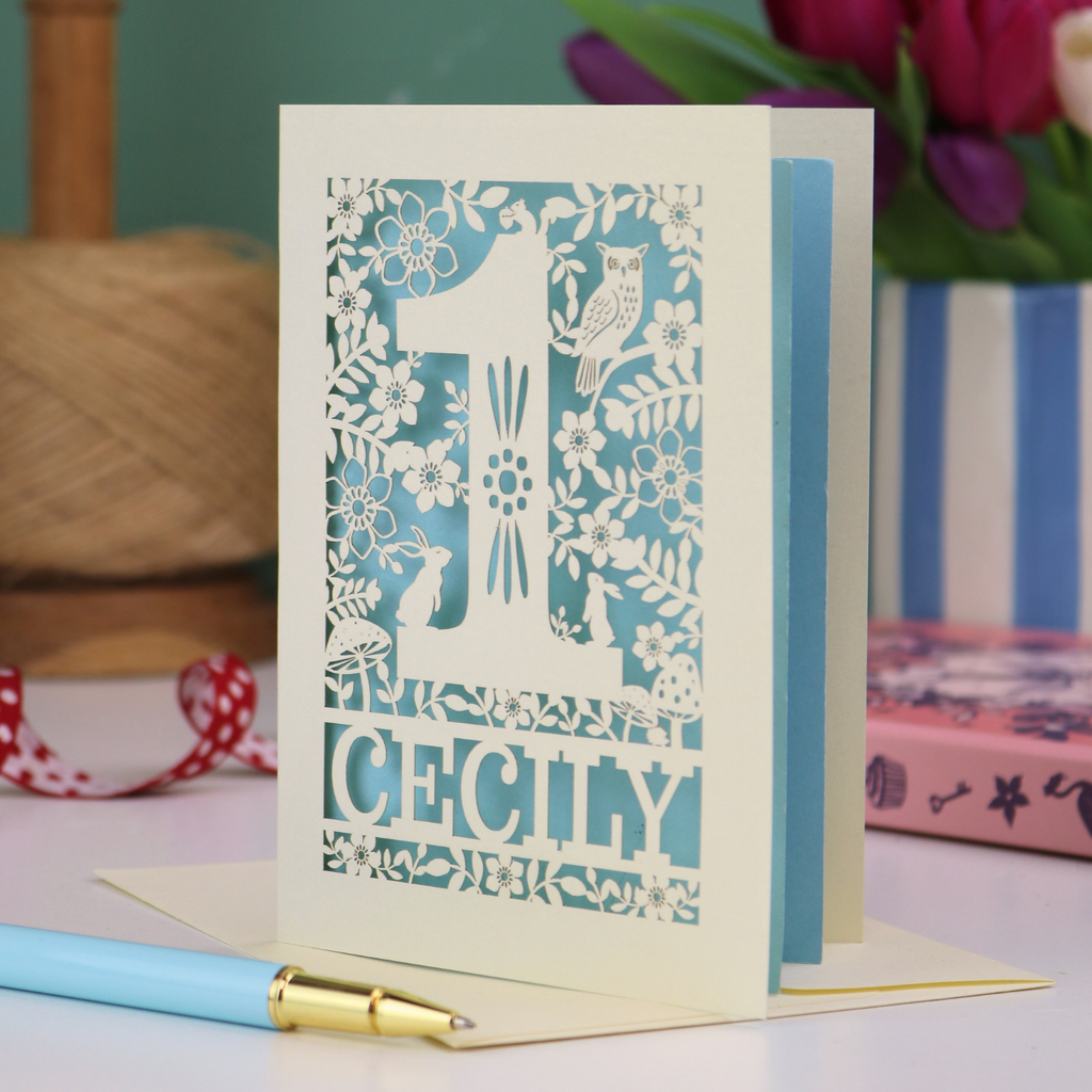 A laser cut 1st birthday card with decorative 1 and woodland design, personalised with a name.