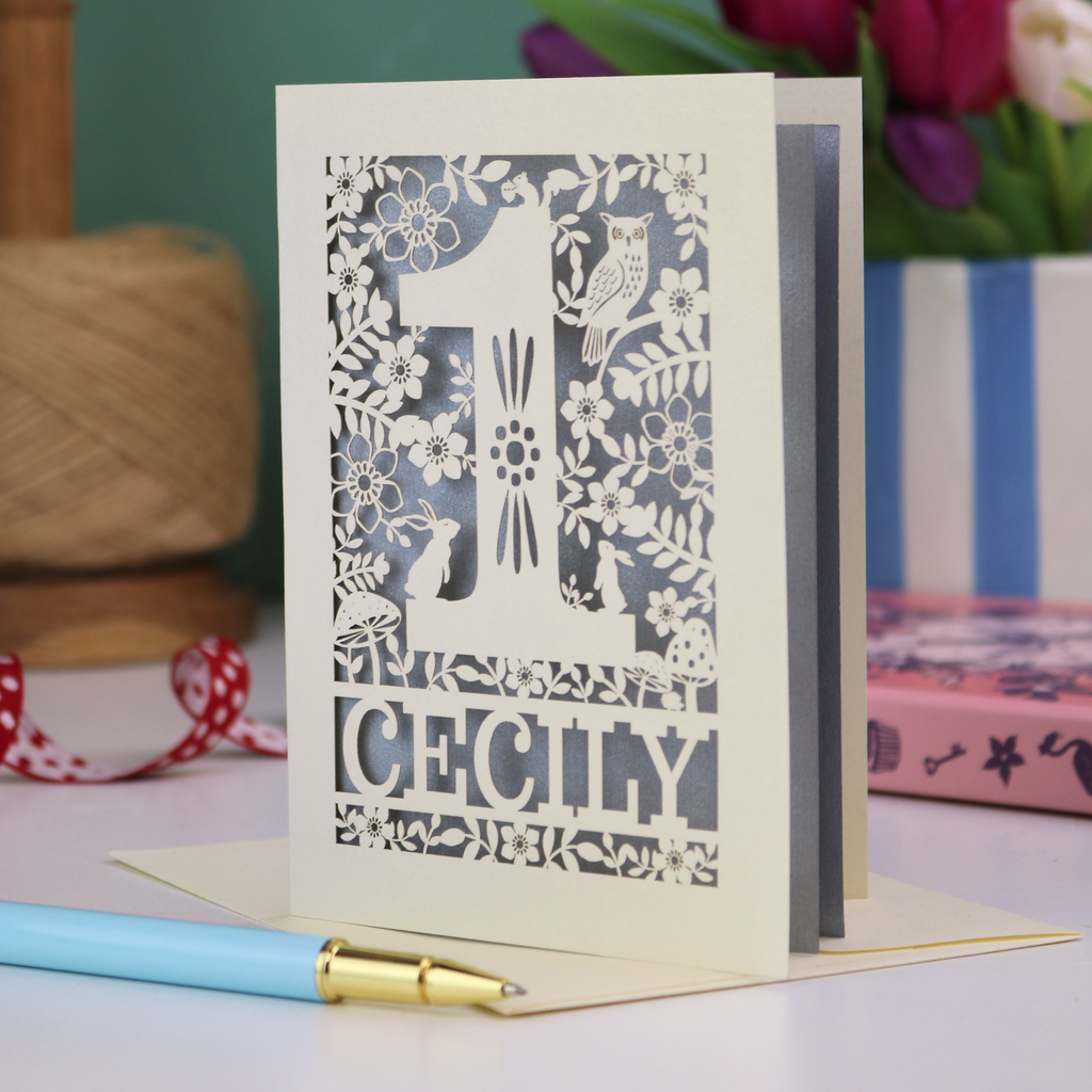 A laser cut 1st birthday card with decorative 1 and woodland design including owl, bunnies, squirrel and hedgehog, personalised with a name.