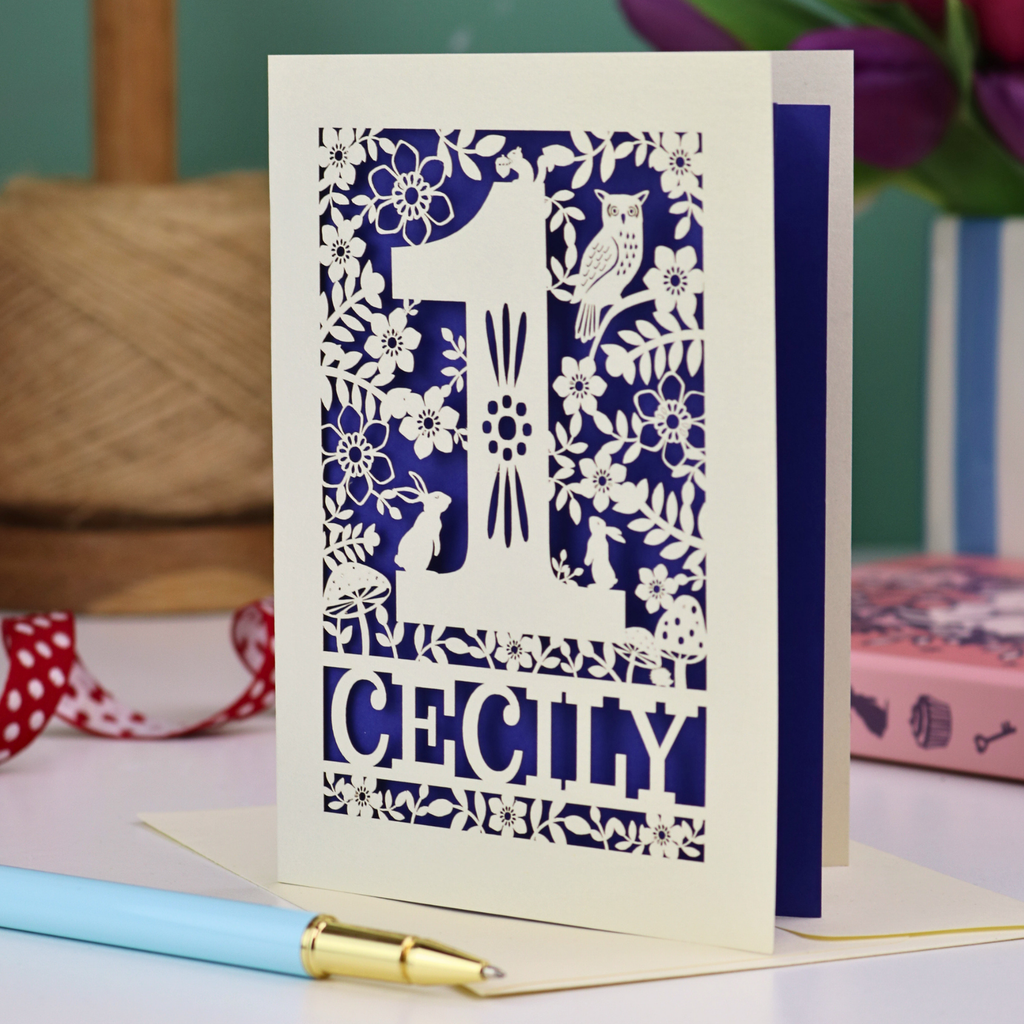 A laser cut 1st birthday card with decorative 1 and woodland design including owl, bunnies, squirrel and hedgehog, personalised with a name.