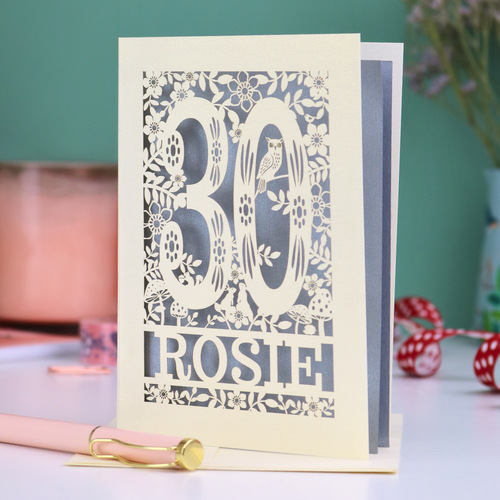 Laser-cut 30th birthday card with '30' and 'Rosie' on a table with decorative items.
