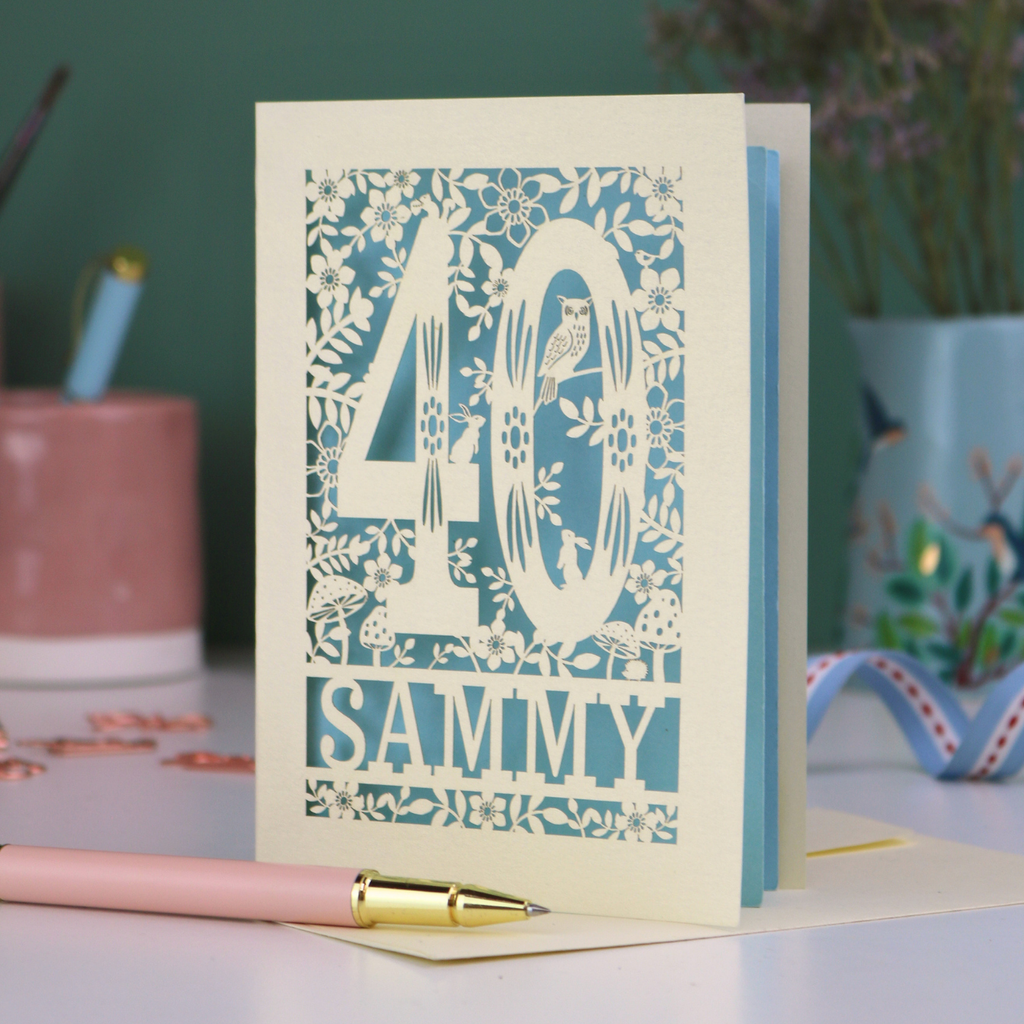 A laser cut 40th birthday card with woodland design, decorative number '40 and personalised with a name, 'Sammy', on a white desk with pen and stationery items