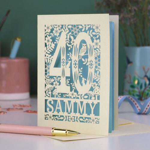 A laser cut 40th birthday card with woodland design, decorative number '40 and personalised with a name, 'Sammy', on a white desk with pen and stationery items