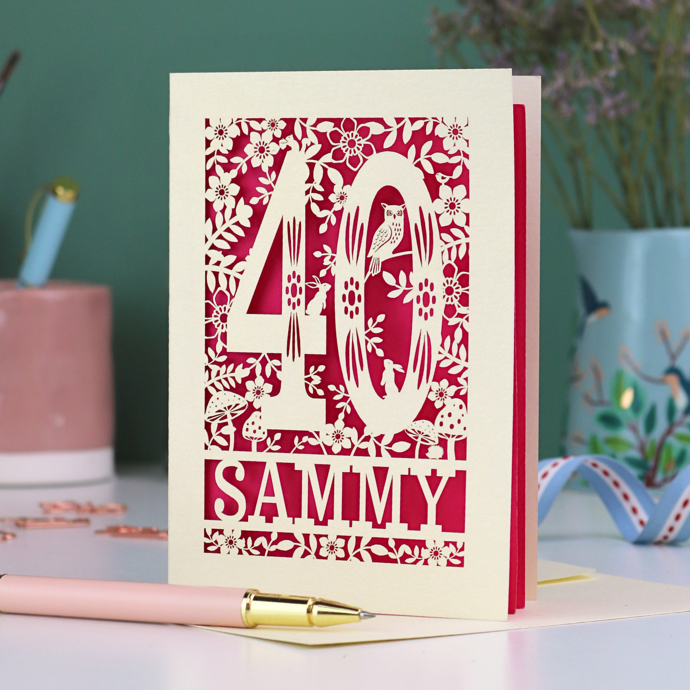 A laser cut 40th birthday card with woodland design, decorative number '40 and personalised with a name, 'Sammy', on a white desk with pen and stationery items