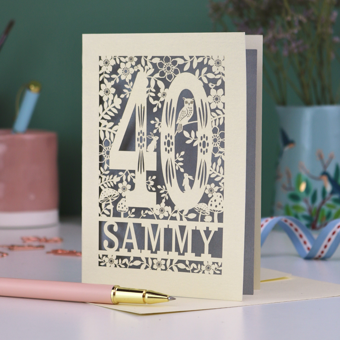 A laser cut 40th birthday card with woodland design, decorative number '40 and personalised with a name, 'Sammy', on a white desk with pen and stationery items