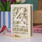Decorative 4th Birthday card with floral and animal laser cut patterns, personalised with the name 'Harley', on a table with stationery items.